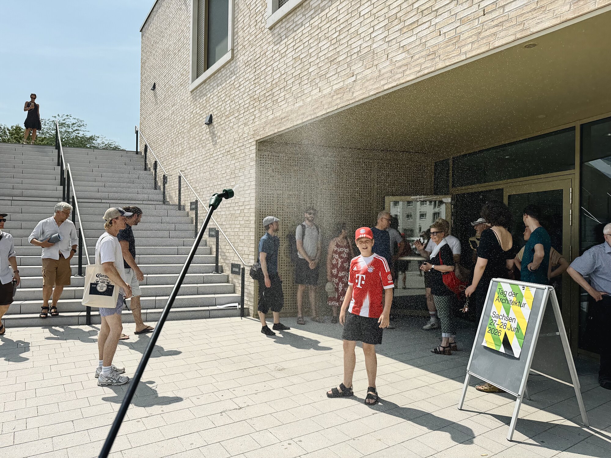 Vor einem modernen Gebäude mit heller Klinkerfassade stehen mehrere Menschen auf einem gepflasterten Platz. Einige Personen tragen Sommerkleidung und Sonnenhüte. Im Vordergrund steht ein Kind mit rotem Trikot, roter Kappe und Brille, das von einem feinen Wassernebel besprüht wird. Links im Bild ist ein schwarzes Rohr oder Gestänge zu sehen, aus dem der Wassernebel austritt. Rechts neben dem Eingang steht ein Aufsteller mit gelbem Plakat. Im Hintergrund führen breite Treppenstufen nach oben, auf denen weitere Menschen gehen.