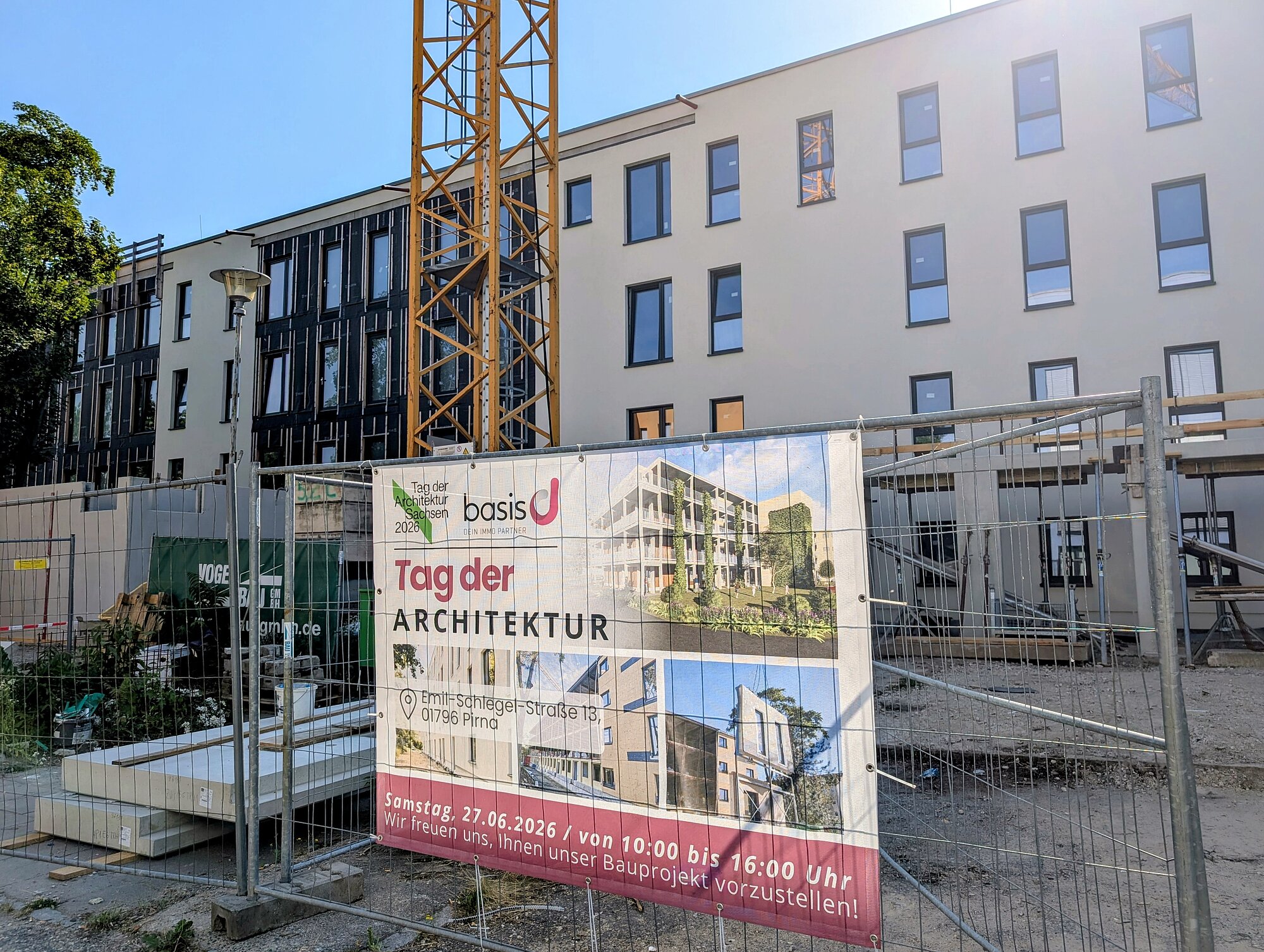 Ein Bauzaun mit einem großen Banner steht vor einem modernen, mehrstöckigen Wohngebäude. Das Banner zeigt eine Visualisierung des Bauprojekts, mehrere Logos und den Schriftzug 'Tag der Architektur'. Im Hintergrund ist das Gebäude mit hellen Fassaden und vielen Fenstern zu sehen. Der Bereich vor dem Gebäude ist eine Baustelle mit Bauzäunen, Baumaterialien und einem Kran. Die Szene ist bei Tageslicht aufgenommen.