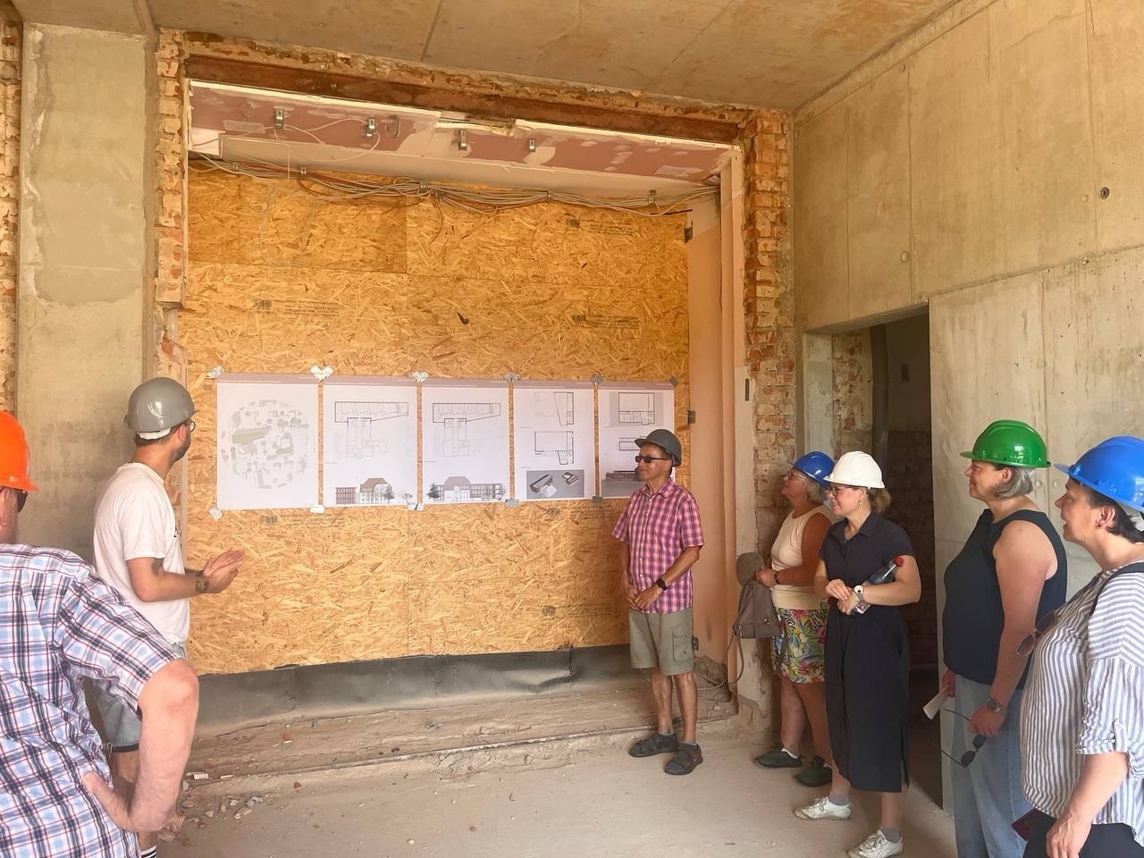 Eine Gruppe von Menschen mit Schutzhelmen steht in einem Rohbau vor einer Spanplattenwand. An der Wand sind mehrere Baupläne nebeneinander befestigt. Eine Person links im Bild zeigt mit der Hand auf die Pläne, während die anderen aufmerksam zuhören. Die Szene spielt in einem unfertigen Innenraum mit unverputzten Wänden und offenliegenden Leitungen.